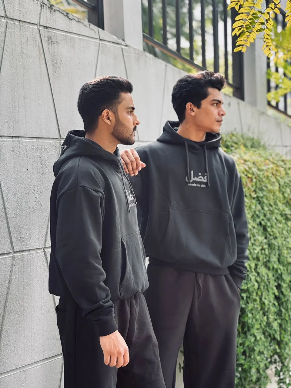 Two models in Obsidian black FAZAL hoodie sets against a concrete wall